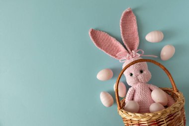 Pastel paskalya yumurtalarıyla çevrili pembe örülü tavşan bebeğiyle örülmüş sepet. Yumuşak mavi arka planda.