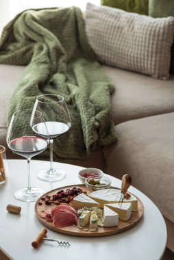 Çeşit çeşit peynirli ahşap tabaklar, kurutulmuş etler, kurutulmuş meyveler, fındık ve zeytinler, yanında iki bardak kırmızı şarap ve kanepede yanan mumlar.