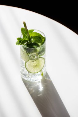 Mojito kokteyli, limon dilimleri, taze nane yaprakları ve buz küpleri, beyaz masadaki çizgili kamışla tamamlanmış.