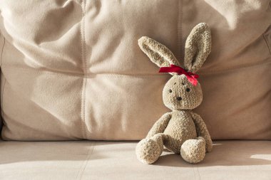 Kırmızı kurdeleli pofuduk tavşancık bej koltukta oturuyor.