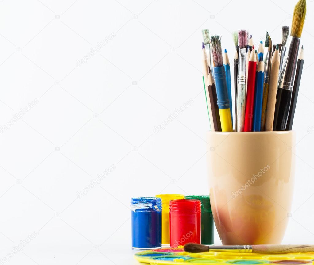 Paint brushes and pencils in mug and colorful paints on white