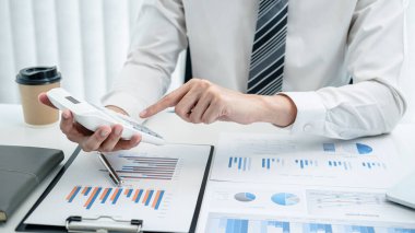 İş adamı yatırım verilerini kontrol etmek için hesap makinesine basar, kitaptaki çeşitli grafiklerden gelen verileri inceler