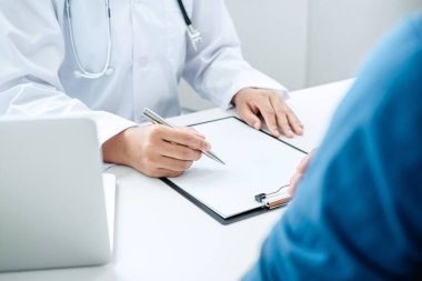 Doktor ve hasta klinikte bir şey tartışıyorlar, hasta masada oturuyor ve ellerini masaya koyuyor, doktor kalemi panoya yazmak için tutuyor.