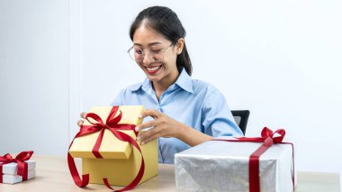 Genç Asyalı bir kadın kırmızı kurdeleli bir hediye kutusu açıyor ve ofis çalışma alanındaki Noel kutlamasının tadını çıkarmak için gülümsüyor.