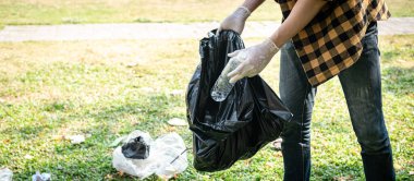 Eldivenli gönüllü adam plastik şişeyi plastik siyah torbaya koyup çevresel aktiviteler sırasında çöpleri toplamak için parkı temizleyecek.
