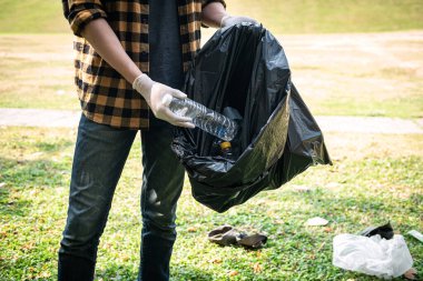 Eldivenli gönüllü adam plastik şişeyi plastik siyah torbaya koyup çevresel aktiviteler sırasında çöpleri toplamak için parkı temizleyecek.