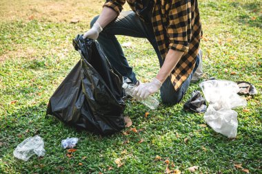 Eldivenli gönüllü bir adam plastik şişe ve plastik atıkları plastik poşete doldurup çevresel aktiviteler sırasında çöpleri toplamak için parkı temizliyor.