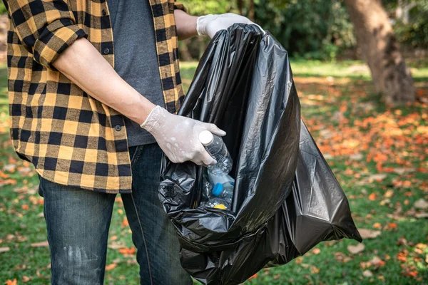 Volunteer man in gloves to picking up plastic bottle into plastic black bag for cleaning the park during environmental activity to collecting garbage