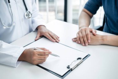 Doktor, klinikteki hastaya belirtileri açıklıyor. Doktor, tedavi detaylarını okuyor ve hastaya reçete yazıyor.