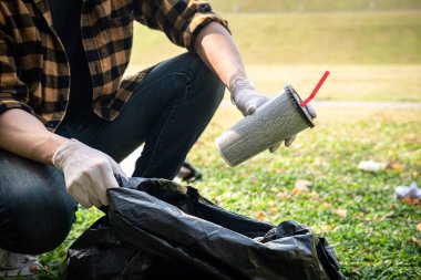 Eldivenli gönüllü bir adam, çevre aktiviteleri sırasında çöpleri toplamak için kahve fincanını plastik siyah bir torbaya koyup parkı temizliyor.