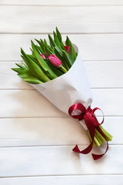 Beyaz ahşap arka planda kırmızı kurdeleli taze kokulu pembe lalelerden oluşan romantik bir buket.