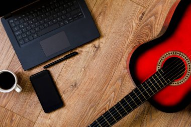 Kırmızı-siyah klasik gitar tahta zeminde duruyor, bilgisayarın, telefonun ve not defterinin yanında.