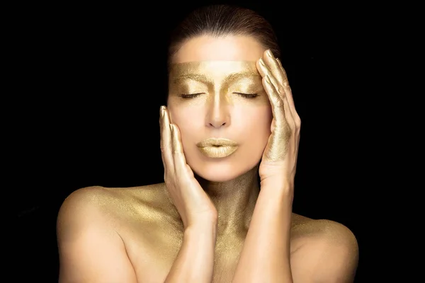 Gold skin girl Isolated on black. Gold based skincare concept. Beauty woman face with gold mask holding a hand on cheek while touching lips with the other with closed eyes in a sensual gesture