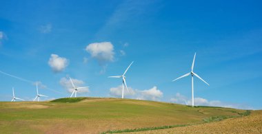 Yel değirmeni enerji dönüşümü. Gökyüzünde beyaz bulutlar olan güneşli bir günde yeşil çimen tarlası ve arka planda dağlar bulunan yel değirmeni türbinleri. Enerji ve ekoloji