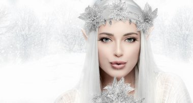 Zarif kış kraliçesi kostümü giymiş güzel bir kadın narin çiçeğe soğuk hava üflüyor ve kış kırsalının arka planındaki kameraya bakıyor.