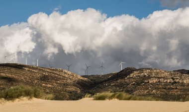 Yenilenebilir veya sürdürülebilir enerji için kıyı rüzgârı çiftliği kumlu bir tropik plaj manzarası ve dağ zeminli kum tepeleri ve yüksek bulut oluşumları olan gökyüzüne karşı rüzgar türbinleri.