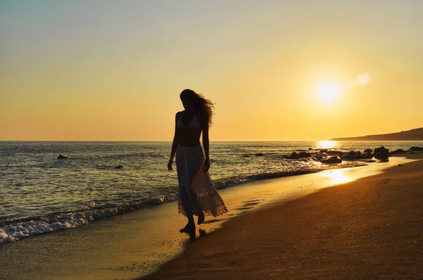 在夕阳的映照下 一个女人在沙滩上走着 背后闪烁着橙色的阳光 在潮湿的沙滩上和海洋上投射出倒影 长长的头发在微风中飘扬 健康的生活和休假概念 — 图库照片