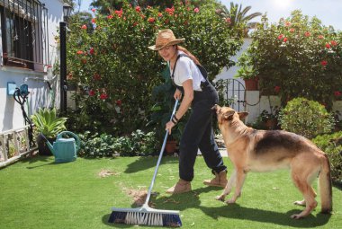 Saman şapkalı genç bir kadın baharda bahçedeki yeşil çimlerden yaprakları süpürüyor. Sadık Alman çoban köpeği tarafından bahçıvanlık ya da mevsim konseptinde izleniyor.