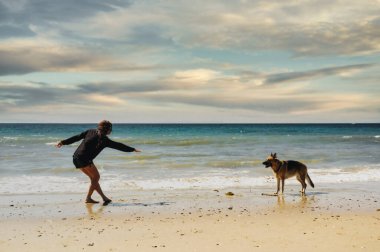 Gün batımında deniz kıyısında köpekle oynayan güzel, genç bir kadın.