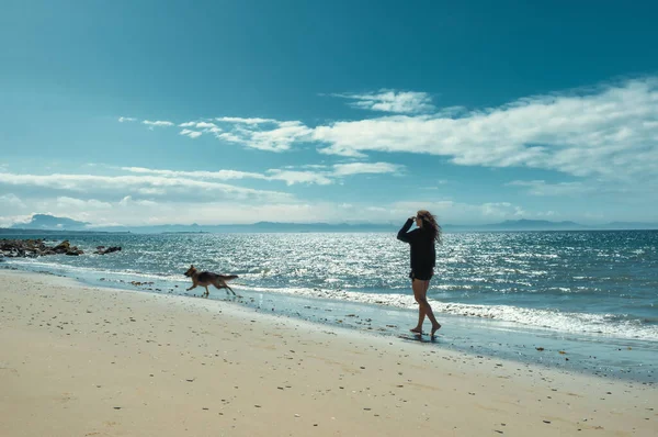 在黄昏的阳光下 与德国牧羊犬在海边的沙滩上玩耍的快乐女人 伴随着轻柔的浪花和闪耀的云彩 — 图库照片