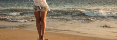 Günbatımında kumlu bir tropik plajda, arkası sörf ve denize dönük bir panorama pankartıyla, modaya uygun kot şortlar giyen genç bir kadının seksi ince bacakları.