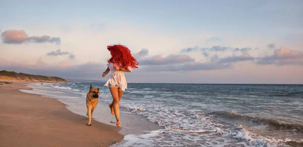 Carefree fit body girl enjoys her holidays running with her German shepherd dog at tropical paradise beach. Happy red-haired woman on travel vacation. Friendship, fun and enjoyment at summer vacation