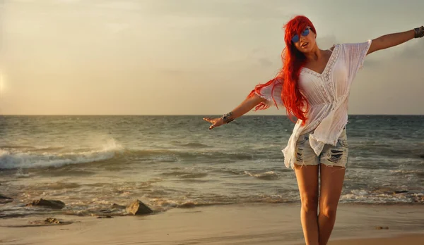 Young woman with bright red dyed hair on a tropical beach at sunset standing with her arms out like an airplane in a travel and summer vacation concept. Relax, fun and enjoy holidays