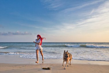 Kumsalda köpeğiyle koşan, yaz havasında mutlu kızıl saçlı bir kadın.