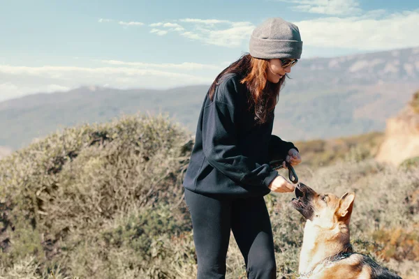 若い女性は彼女のドイツの羊飼いの犬に丘や山の上で一緒に田舎の屋外での訓練中の御馳走を提供します — ストック写真