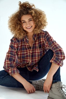 Sarı kıvırcık saçlı, gülümseyen genç kadın pamuklu gömlek giyiyor ve beyaz arka planda oturuyor.