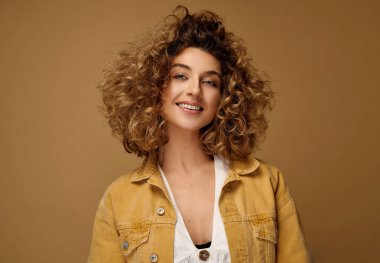 Stüdyoda kahverengi stüdyo arka planında poz veren kıvırcık saçlı mutlu genç kadın. Sarışın, afro saçlı, gülümseyen kadın. 