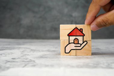 Elinde kırmızı çatılı bir evin resmi olan tahta küpler emlak sigortasını, ev yatırımını, ipoteği veya emlak bakımını sembolize ediyor. Konut güvenliği, mali koruma ve aile yatırımı kavramı.