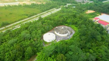 Yoğun yeşil ormanlarla çevrili su depolama tanklarının hava görüntüsü, bir yol ve tesis alanının yakınında, doğal manzarayla harmanlanmış altyapıyı gösteriyor..