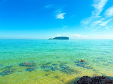 Pantai Bukit Keluang 'ın berrak güneşli manzarası turkuaz suları, hafif dalgaları ve ufukta görünen bir adası var..