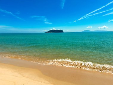 Pantai Bukit Keluang 'ın berrak güneşli manzarası turkuaz suları, hafif dalgaları ve ufukta görünen bir adası var..