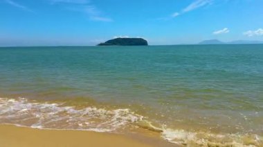 Yumuşak dalgalar açık mavi gökyüzünün altında uzak bir adayla kumlu bir sahile vuruyor..