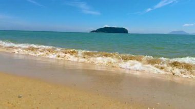 Yumuşak dalgalar açık mavi gökyüzünün altında uzak bir adayla kumlu bir sahile vuruyor..