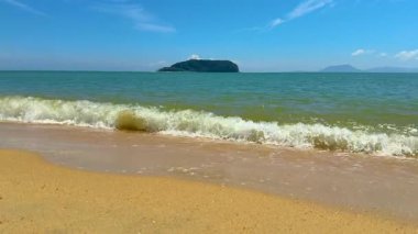 Yumuşak dalgalar açık mavi gökyüzünün altında uzak bir adayla kumlu bir sahile vuruyor..