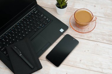 Bilgisayarlı, akıllı telefonlu, not defterli ve beyaz ahşap masadaki kahve fincanı ile en üst düzey çalışma alanı üretkenliği, dijital işi, günlük rutini ve temiz kopya alanı olan profesyonel ofis düzenini temsil ediyor..