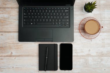 Bilgisayarlı, akıllı telefonlu, not defterli ve beyaz ahşap masadaki kahve fincanı ile en üst düzey çalışma alanı üretkenliği, dijital işi, günlük rutini ve temiz kopya alanı olan profesyonel ofis düzenini temsil ediyor..