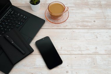 Bilgisayarlı, akıllı telefonlu, not defterli ve beyaz ahşap masadaki kahve fincanı ile en üst düzey çalışma alanı üretkenliği, dijital işi, günlük rutini ve temiz kopya alanı olan profesyonel ofis düzenini temsil ediyor..