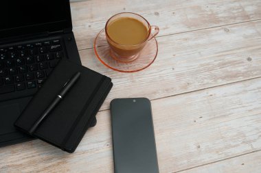 Bilgisayarlı, akıllı telefonlu, not defterli ve beyaz ahşap masadaki kahve fincanı ile en üst düzey çalışma alanı üretkenliği, dijital işi, günlük rutini ve temiz kopya alanı olan profesyonel ofis düzenini temsil ediyor..