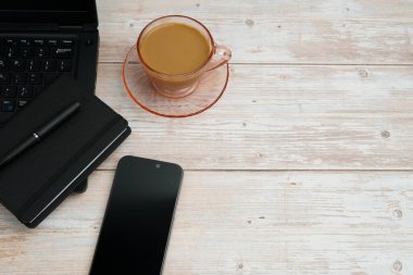 Bilgisayarlı, akıllı telefonlu, not defterli ve beyaz ahşap masadaki kahve fincanı ile en üst düzey çalışma alanı üretkenliği, dijital işi, günlük rutini ve temiz kopya alanı olan profesyonel ofis düzenini temsil ediyor..