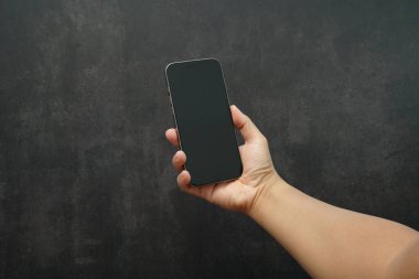 Hand holding smartphone with blank black screen on dark background. Concept of mobile mockup, app presentation, digital marketing, UI UX design, advertising, and technology promotion.