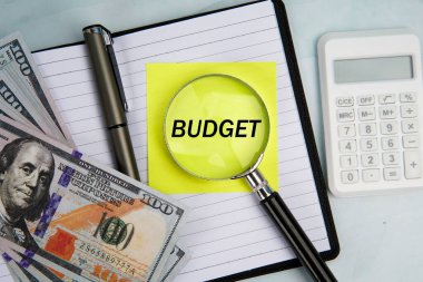 Hesap makinesi, defter, kalem ve Amerikan doları faturalarının yanında bütçe okunurken, mali planlama, gider kontrolü ve para yönetimi kavramını temsil eden yapışkan notlar üzerine büyüteç