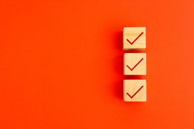 Parlak turuncu arkaplan, kontrol listesi ve görev tamamlama konseptine dikey hizalanmış kırmızı işaretli üç tahta küp.