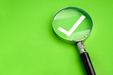 Yeşil arka planda beyaz kontrol işaretini vurgulayan büyüteç, onay, doğrulama ve kalite kontrol kavramını temsil ediyor.