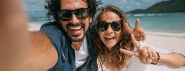 Bir çift aşık tropik bir plajda selfie çekiyor. Adam ve kız denizin arka planındaki telefon kamerasına neşeyle gülümsüyor. Yeni evliler ve yeşil dağları ve bulutlu gökyüzü olan tropik bir göl..