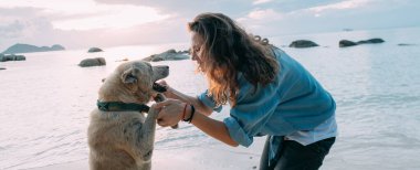 Kız gün batımında sahilde köpekle oynarken eğleniyor. Genç Avrupalı bir kadın deniz ve batan güneşin arka planında evcil bir köpekle yürüyor..