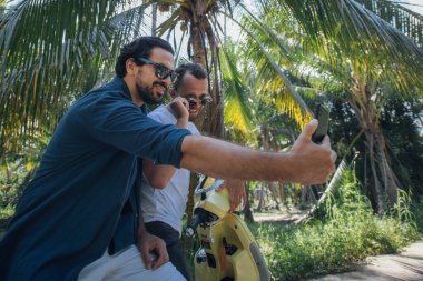 Erkekler tropik bir adada palmiye ağaçlarında motosikletle selfie çekerken eğleniyorlar. Erkek turistler telefonla Palm Grove 'da bisikletle fotoğraf çektiriliyor.